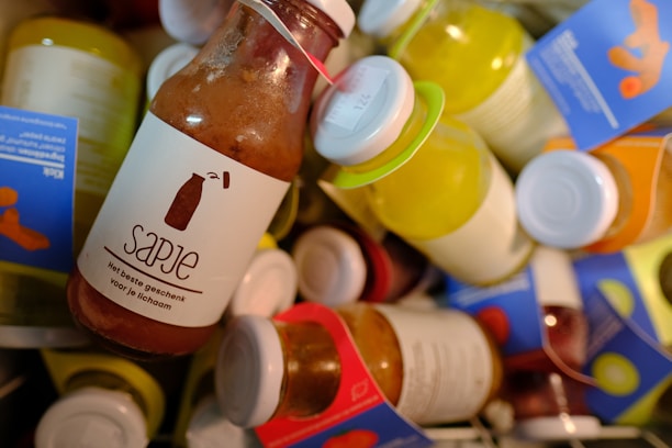 A collection of colorful bottled juices with labels, featuring a prominent brown bottle labeled 'Sapje' in the foreground. The labels are written in Dutch and include phrases related to health. The bottles vary in color with caps in different hues.