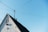 A gabled roof of a house with a rustic appearance. The house exterior is white with a small window featuring closed wooden shutters. There is a birdhouse attached to the wall beneath the window. Overhead, there are multiple power lines connected to a pole on the roof. The sky is clear and blue, creating a peaceful background.