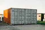 A large shipping container, predominantly gray with the right side illuminated by the sunlight, casting a warm glow that gives it an orange tint, is situated outdoors on a gravel surface. In the background, there are modern buildings and a patch of green grass alongside a small tree or shrub.
