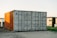 A large shipping container, predominantly gray with the right side illuminated by the sunlight, casting a warm glow that gives it an orange tint, is situated outdoors on a gravel surface. In the background, there are modern buildings and a patch of green grass alongside a small tree or shrub.