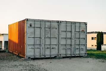 A large shipping container, predominantly gray with the right side illuminated by the sunlight, casting a warm glow that gives it an orange tint, is situated outdoors on a gravel surface. In the background, there are modern buildings and a patch of green grass alongside a small tree or shrub.