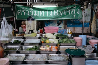 A street food stall displaying a variety of fresh vegetables, herbs, and other ingredients in metal trays. An R2-D2 model is prominently placed on the counter. The backdrop features a green sign with text written in Thai. There are stacks of pink and blue plastic bowls and some hanging plastic bags.