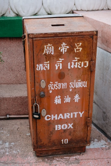 A rusty, rectangular metal box labeled 'CHARITY BOX' with multiple languages inscribed on its surface. It stands on a concrete surface next to a white decorative element, with a padlock securing it.