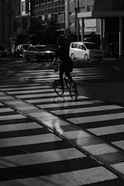 A cyclist is riding across a pedestrian crosswalk on a city street. The scene is in black and white, with vehicles visible in the background. Shadows are cast dramatically across the scene, suggesting evening or early morning light.