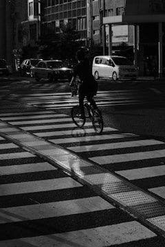 A cyclist is riding across a pedestrian crosswalk on a city street. The scene is in black and white, with vehicles visible in the background. Shadows are cast dramatically across the scene, suggesting evening or early morning light.