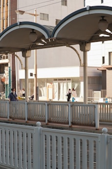 A modern train platform with people walking along the walkway. The platform has a covered, arched structure with lighting fixtures beneath. The background shows urban buildings and some storefronts. Rail tracks run in front of the platform, bordered by metal railings.