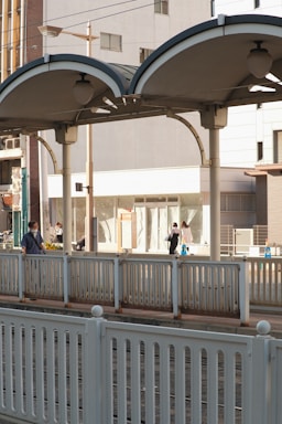 A modern train platform with people walking along the walkway. The platform has a covered, arched structure with lighting fixtures beneath. The background shows urban buildings and some storefronts. Rail tracks run in front of the platform, bordered by metal railings.