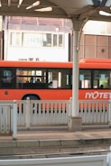 A vibrant orange bus is parked or in motion near a station platform with white railings. The bus has sleek, modern windows, and there are several passengers visible inside. In the background is an urban setting with a building displaying a sign reading 'Enchanté'. The architecture includes varying shades of brown and white.