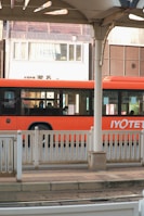 A vibrant orange bus is parked or in motion near a station platform with white railings. The bus has sleek, modern windows, and there are several passengers visible inside. In the background is an urban setting with a building displaying a sign reading 'Enchanté'. The architecture includes varying shades of brown and white.