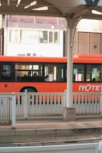 A vibrant orange bus is parked or in motion near a station platform with white railings. The bus has sleek, modern windows, and there are several passengers visible inside. In the background is an urban setting with a building displaying a sign reading 'Enchanté'. The architecture includes varying shades of brown and white.