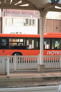 A vibrant orange bus is parked or in motion near a station platform with white railings. The bus has sleek, modern windows, and there are several passengers visible inside. In the background is an urban setting with a building displaying a sign reading 'Enchanté'. The architecture includes varying shades of brown and white.