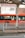 A vibrant orange bus is parked or in motion near a station platform with white railings. The bus has sleek, modern windows, and there are several passengers visible inside. In the background is an urban setting with a building displaying a sign reading 'Enchanté'. The architecture includes varying shades of brown and white.