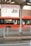A vibrant orange bus is parked or in motion near a station platform with white railings. The bus has sleek, modern windows, and there are several passengers visible inside. In the background is an urban setting with a building displaying a sign reading 'Enchanté'. The architecture includes varying shades of brown and white.
