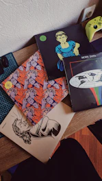 Several vinyl record covers are spread out on a wooden surface. One cover features an illustration of a person with a serious expression, another has a colorful pattern with multiple faces, and a third displays a globe with the words 'MORE DATA'. A bright green game controller is partially visible on the side.
