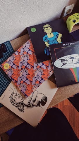 Several vinyl record covers are spread out on a wooden surface. One cover features an illustration of a person with a serious expression, another has a colorful pattern with multiple faces, and a third displays a globe with the words 'MORE DATA'. A bright green game controller is partially visible on the side.