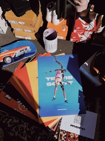 A collection of colorful posters with a prominent one depicting a person in an artistic pose. The surroundings include various shoe boxes, some branded with recognizable logos, and a skateboard featuring a car design. A patterned carpet is partially visible, and an upright cylinder with abstract designs and text is placed among the items.