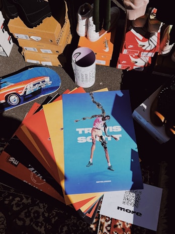 A collection of colorful posters with a prominent one depicting a person in an artistic pose. The surroundings include various shoe boxes, some branded with recognizable logos, and a skateboard featuring a car design. A patterned carpet is partially visible, and an upright cylinder with abstract designs and text is placed among the items.
