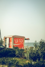 A bright orange kiosk is situated near lush greenery with a view of the sea in the background. The kiosk has Arabic script written on it and is partially obscured by plants. The scene suggests a coastal or beachside location during the daytime.