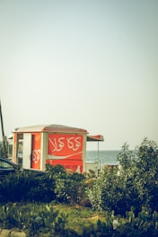 A bright orange kiosk is situated near lush greenery with a view of the sea in the background. The kiosk has Arabic script written on it and is partially obscured by plants. The scene suggests a coastal or beachside location during the daytime.