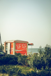 A bright orange kiosk is situated near lush greenery with a view of the sea in the background. The kiosk has Arabic script written on it and is partially obscured by plants. The scene suggests a coastal or beachside location during the daytime.