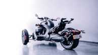 A sleek, modern three-wheeled motorcycle is parked on a smooth, reflective floor. The design features a matte black finish with angular lines and accents in orange. Two large rear wheels and a single front wheel are visible, along with handlebars and side mirrors. The bike is positioned in a clean, minimalist setting with neutral lighting that highlights its futuristic aesthetics.