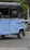 A blue auto rickshaw with a black canopy is parked on a cobblestone street. The vehicle shows some wear and is missing some body parts, revealing part of its interior. The pavement contains a few scattered brown leaves, and in the background, there is greenery with some vegetation visible.