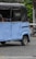 A blue auto rickshaw with a black canopy is parked on a cobblestone street. The vehicle shows some wear and is missing some body parts, revealing part of its interior. The pavement contains a few scattered brown leaves, and in the background, there is greenery with some vegetation visible.