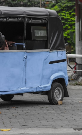 A blue auto rickshaw with a black canopy is parked on a cobblestone street. The vehicle shows some wear and is missing some body parts, revealing part of its interior. The pavement contains a few scattered brown leaves, and in the background, there is greenery with some vegetation visible.