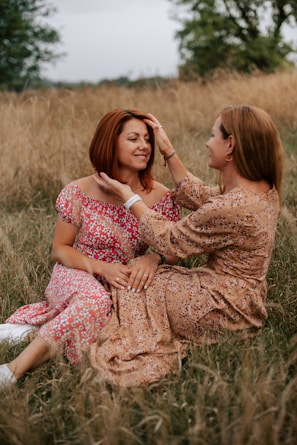 Two women are sitting in a field of tall grass, engaging in a tender moment. They are both wearing floral dresses. One woman is gently holding the other's face, and they are both smiling warmly at each other. The scene is set outdoors with greenery and an overcast sky in the background.