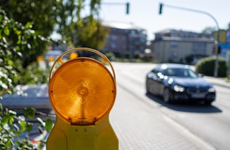 A road with a black car driving while a yellow caution light is prominently featured in the foreground. The background includes blurred buildings and green foliage.