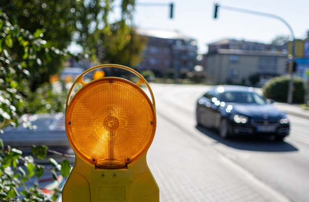 A road with a black car driving while a yellow caution light is prominently featured in the foreground. The background includes blurred buildings and green foliage.