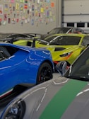 Several high-end sports cars are parked inside a garage with a wall covered in various colorful art pieces. The cars include a blue car with black rims, a bright yellow car with aggressive styling, and a gray car with green stripes.