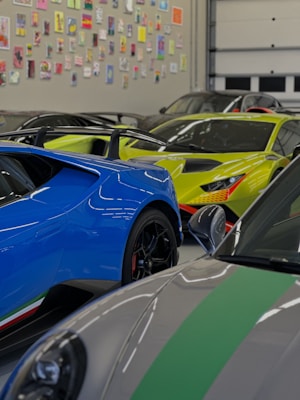 Several high-end sports cars are parked inside a garage with a wall covered in various colorful art pieces. The cars include a blue car with black rims, a bright yellow car with aggressive styling, and a gray car with green stripes.