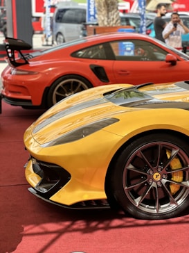Two high-performance sports cars are parked close together on a red carpet. The foreground features a yellow Ferrari with sleek lines and distinctive wheel design, while a red Porsche with a prominent rear wing is positioned behind it. The background includes blurred figures and a transport van, indicating an event or display setting.