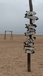 A weathered wooden post on a sandy beach features multiple arrow-shaped signs pointing in different directions. The signs display words like 'VERTIGO', 'SURFSHOP', 'KITE SCHOOL', 'SUP', 'BIKINIS', 'ALOHA', 'KITES', 'FUN', 'SUNSCREEN', and 'BUGGY'. In the background, a simple wooden swing set is situated further down the beach. The sky is overcast and the overall atmosphere appears calm and serene.