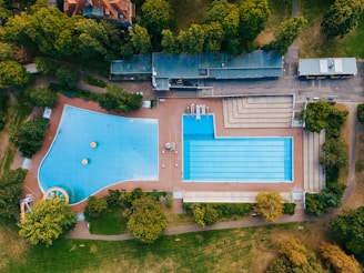 Aerial view of a large outdoor swimming pool complex surrounded by trees. The complex includes two distinct pools, one with lanes marked for lap swimming and another irregularly shaped pool with a water slide and floating buoy markers. The surrounding area features greenery and some buildings, likely facilities related to the pool.