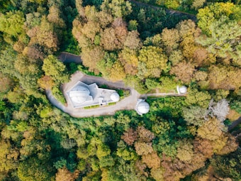 Aerial view of an observatory surrounded by dense, lush forest. The structure includes a main building with a few dome-topped extensions, nestled among vibrant green foliage and winding pathways.