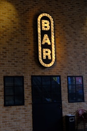 An illuminated vertical sign with the word 'BAR' in bold, surrounded by light bulbs, is mounted on a brick wall. Below the sign are three large, dark-framed glass windows. A dark door with similar framing matches the windows, and a red chair is partially visible at the bottom right.