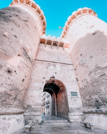 A historic stone gateway with two large cylindrical towers, weathered and textured by time. The gateway features an arched entrance that leads to a street lined with buildings. The sky above is clear and blue, and a small flag is visible atop the structure.