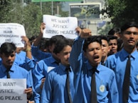A group of young men wearing blue shirts and black ties are participating in a protest. Some are holding up signs with messages demanding justice and peace. The mood is assertive and determined.