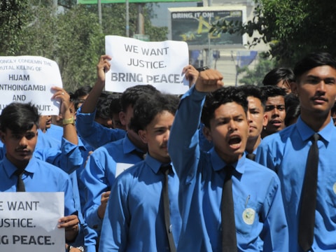 A group of young men wearing blue shirts and black ties are participating in a protest. Some are holding up signs with messages demanding justice and peace. The mood is assertive and determined.