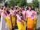 A group of young women in pink shirts and yellow skirts stand together, with one of them prominently shouting or chanting. They are outdoors, possibly during a protest or demonstration. Some are wearing face masks, and many have signs or badges around their necks. In the background, uniformed personnel can be seen.