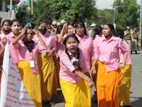 A group of young women in pink shirts and yellow skirts stand together, with one of them prominently shouting or chanting. They are outdoors, possibly during a protest or demonstration. Some are wearing face masks, and many have signs or badges around their necks. In the background, uniformed personnel can be seen.