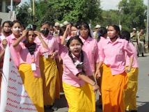 A group of young women in pink shirts and yellow skirts stand together, with one of them prominently shouting or chanting. They are outdoors, possibly during a protest or demonstration. Some are wearing face masks, and many have signs or badges around their necks. In the background, uniformed personnel can be seen.