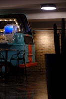 A retro van with a turquoise and orange color scheme is repurposed as a bar setup in an indoor space. The interior includes shelves stocked with bottles and is illuminated with a series of lights. Stylish black chairs are positioned nearby on a dark reflective floor. The backdrop features a brick wall adding a rustic touch, and a ceiling light provides additional illumination.