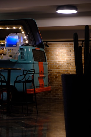 A retro van with a turquoise and orange color scheme is repurposed as a bar setup in an indoor space. The interior includes shelves stocked with bottles and is illuminated with a series of lights. Stylish black chairs are positioned nearby on a dark reflective floor. The backdrop features a brick wall adding a rustic touch, and a ceiling light provides additional illumination.