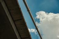 Concrete bridge structure seen from below, against a bright blue sky with soft, white clouds. A large pipe or architectural beam spans the view diagonally, providing a sense of scale and industrial design.