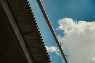 Concrete bridge structure seen from below, against a bright blue sky with soft, white clouds. A large pipe or architectural beam spans the view diagonally, providing a sense of scale and industrial design.