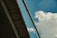 Concrete bridge structure seen from below, against a bright blue sky with soft, white clouds. A large pipe or architectural beam spans the view diagonally, providing a sense of scale and industrial design.