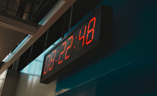 A digital clock displaying the time 08:22:48 with bright red LED numbers, mounted on a blue wall in an indoor setting. The surrounding architecture features modern metallic and glass elements, with overhead panels partially visible.